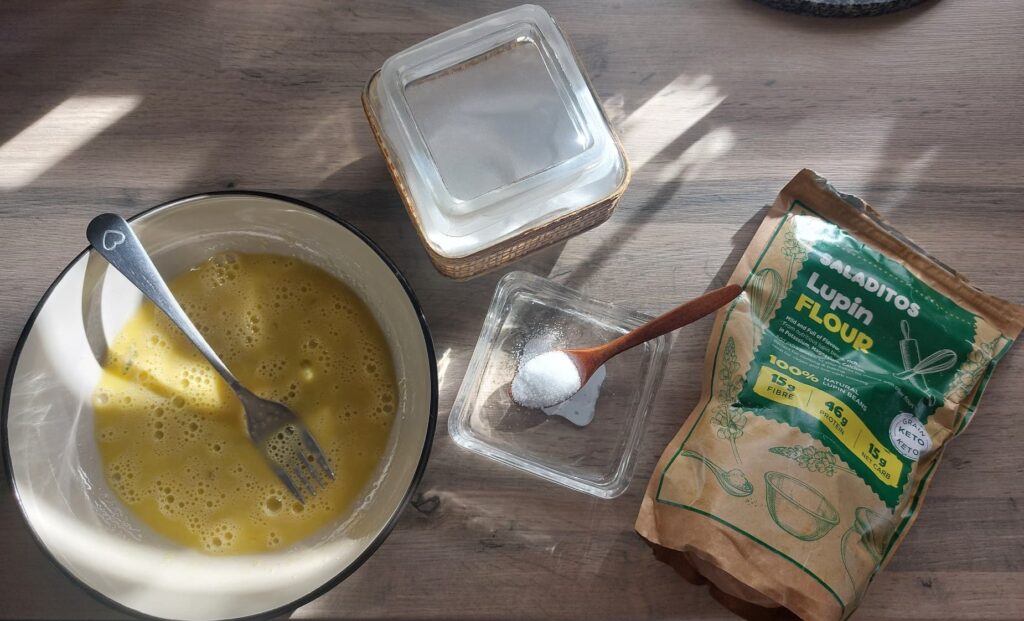 Příprava ingrediencí - Mísa s rozšlehanými vejci a vidličkou položená na kuchyňské lince, vedle ní nádoba s cukrem, malá miska s odměřeným kypřicím práškem a balení lupinové mouky.