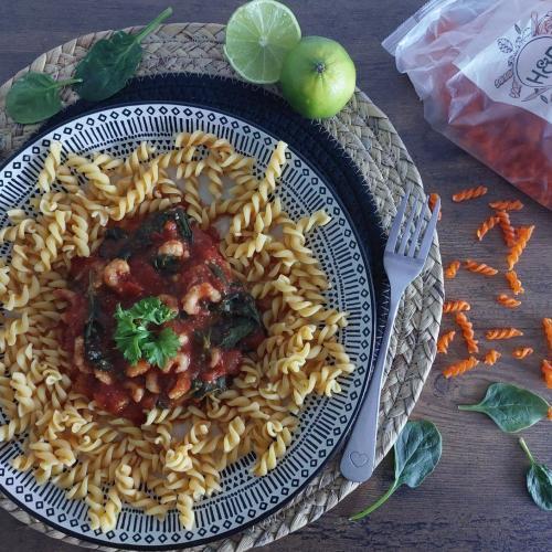 Krevety s rajčaty a špenátem podávané s čočkovými fusilli na modro-bílém talíři, vedle limetka a vidlička