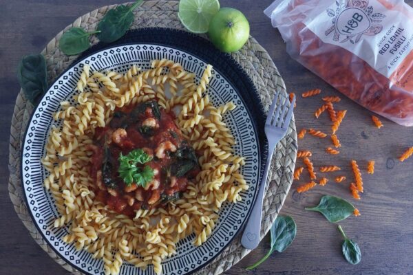 Krevety s rajčaty a špenátem podávané s čočkovými fusilli na modro-bílém talíři, vedle limetka a vidlička