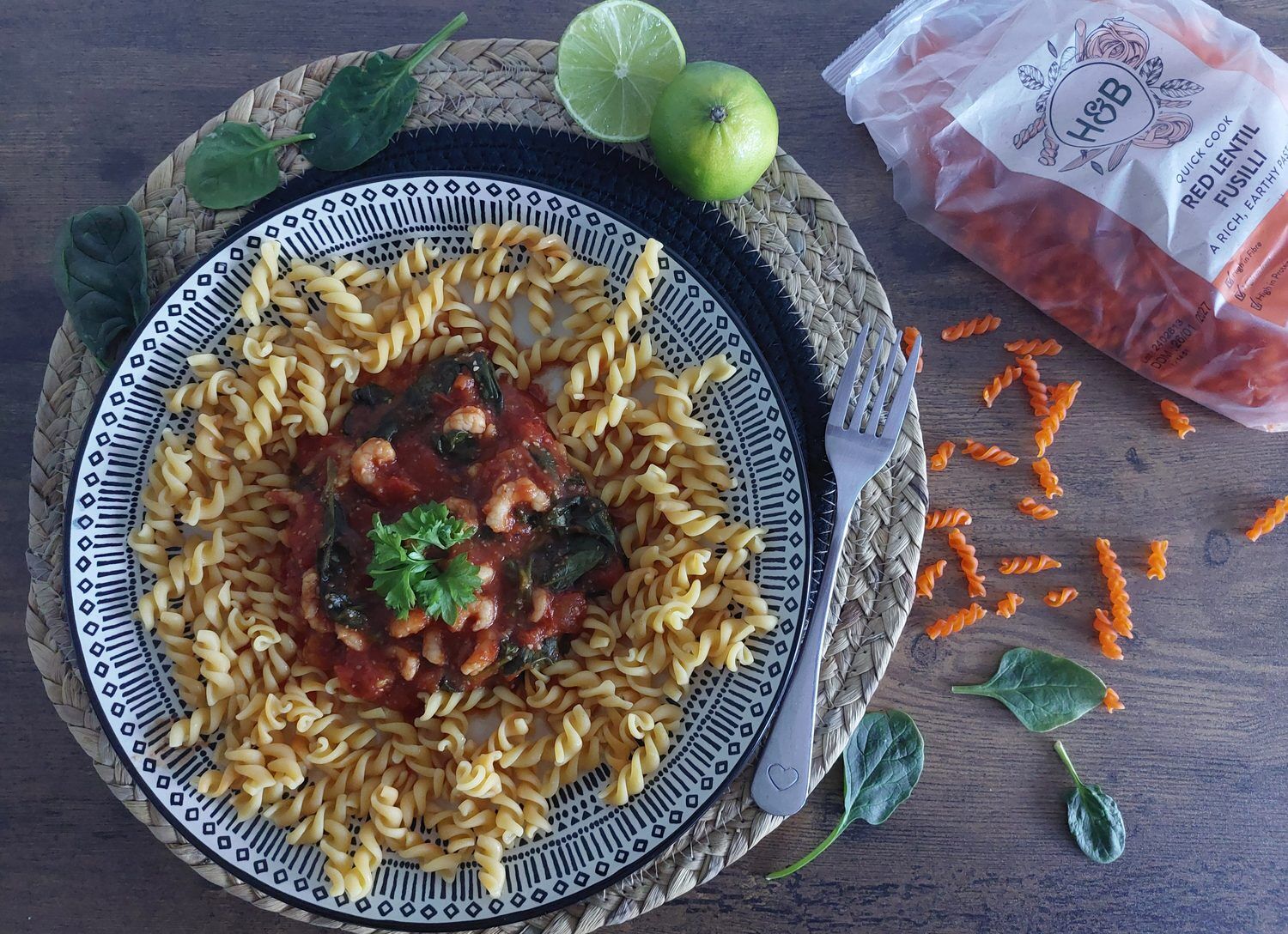 Krevety s rajčaty a špenátem podávané s čočkovými fusilli na modro-bílém talíři, vedle limetka a vidlička